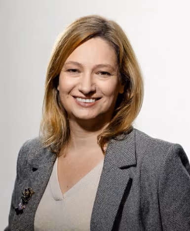 Femme souriante aux cheveux blonds portant un blazer gris et un pull beige, posant devant un fond clair.