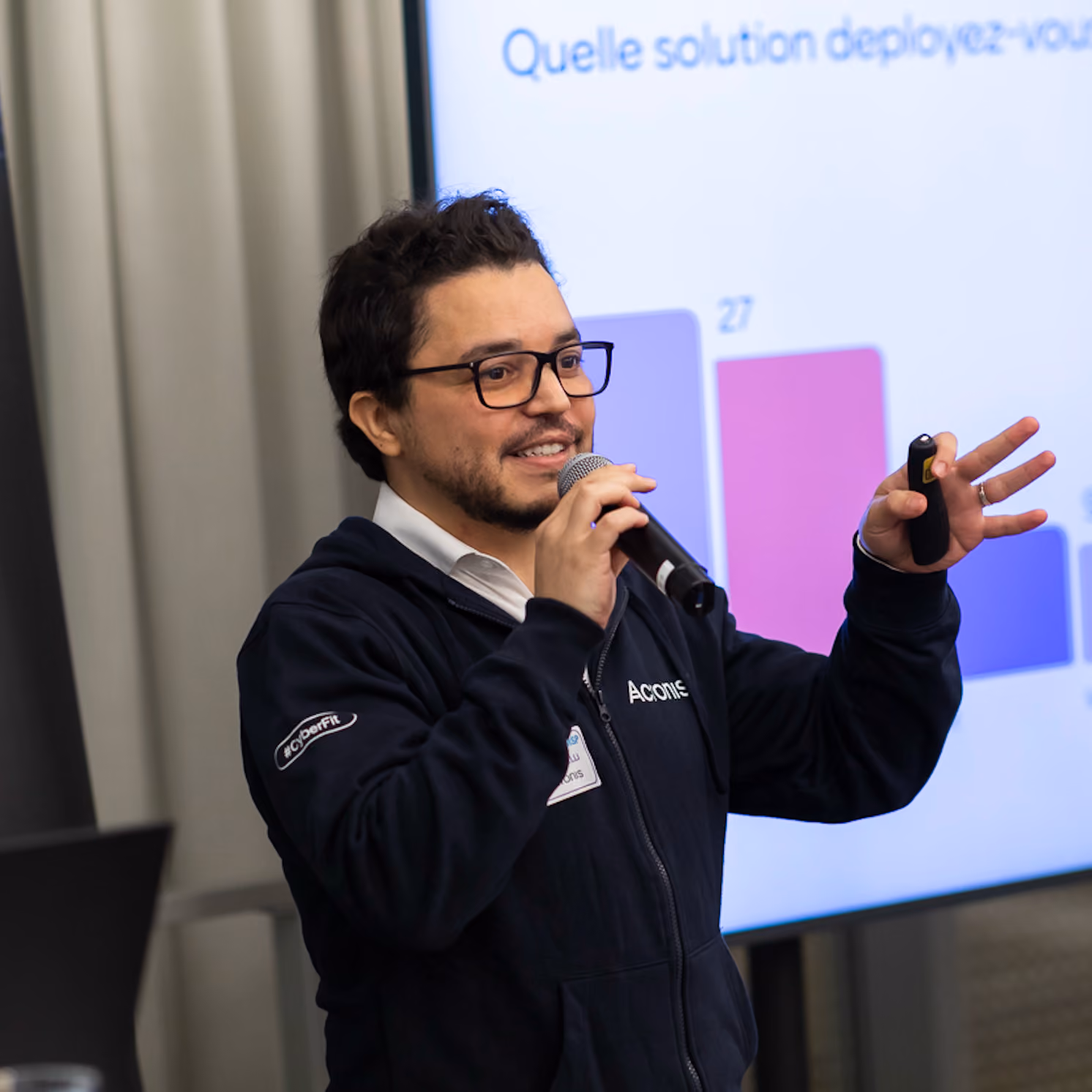 Un homme portant des lunettes et une veste Acronis parle avec un micro devant un écran affichant un graphique coloré et un texte en français.