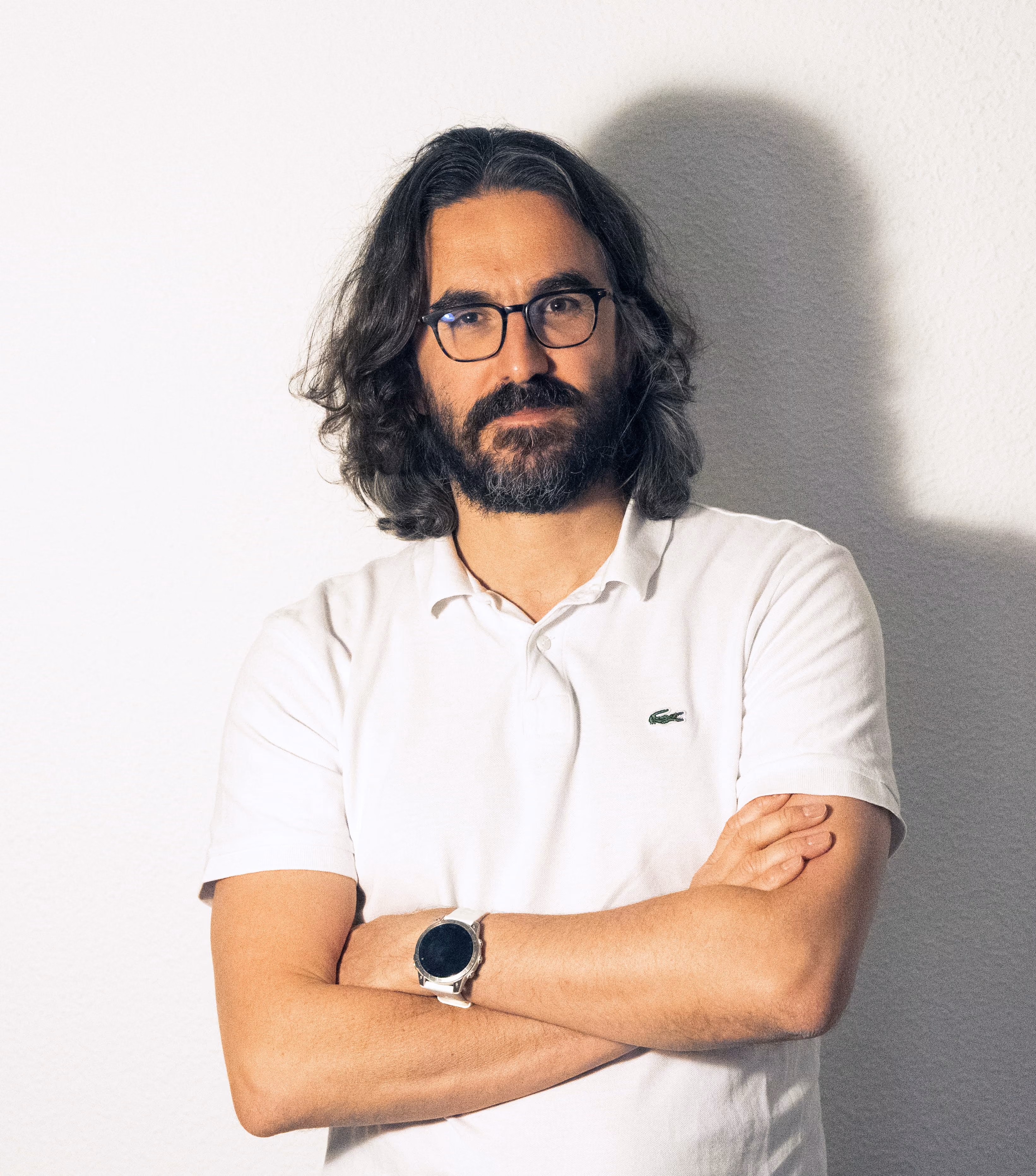 Homme avec cheveux longs, barbe et lunettes portant un polo blanc, bras croisés, devant un mur clair.