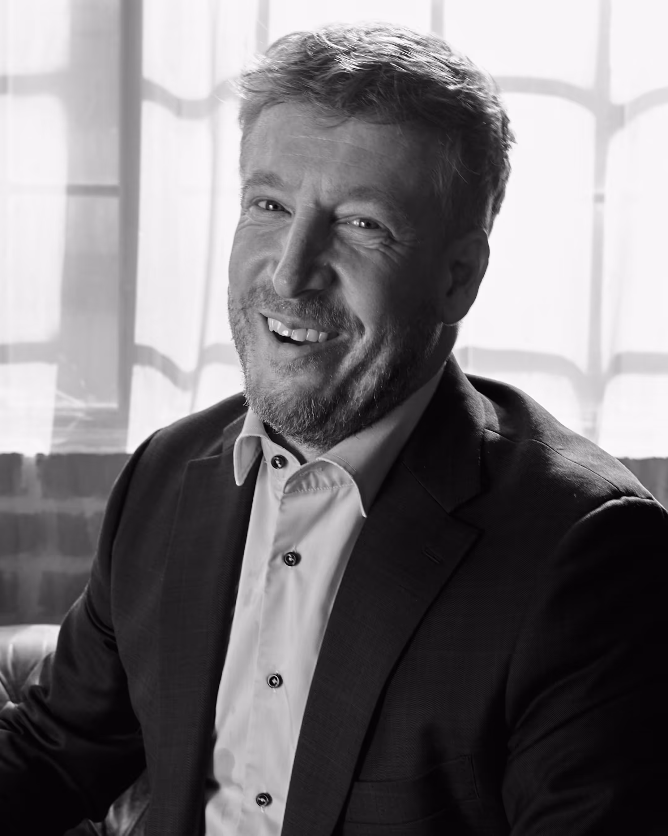 Smiling man with beard wearing a suit jacket and a button-up shirt sitting indoors in front of a window with sheer curtains.