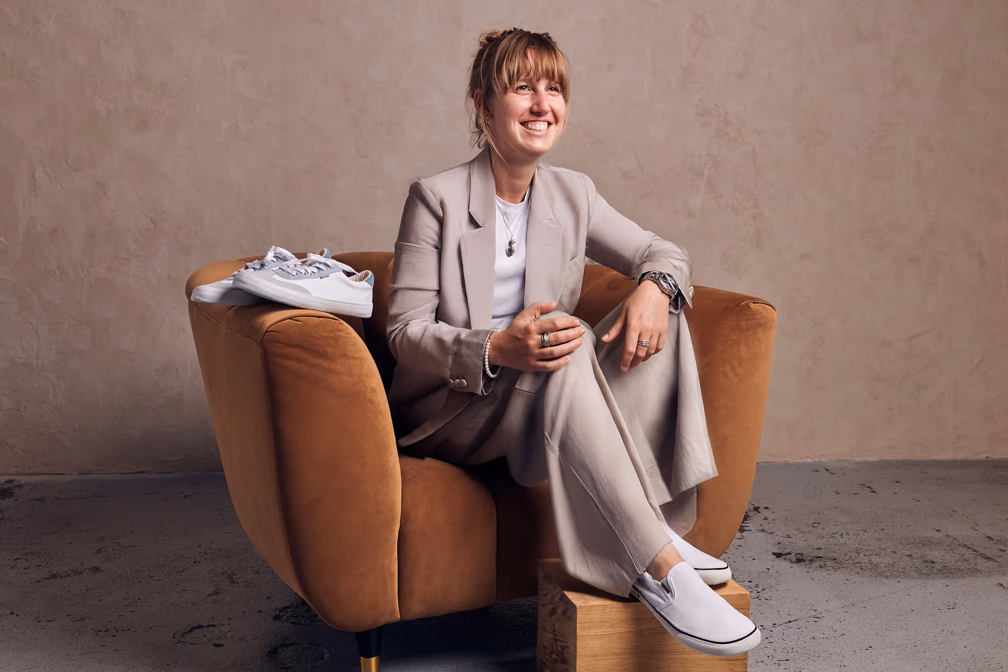 Smiling woman in light gray suit sitting on a brown armchair with white slip-on shoes on a wooden block and white sneakers on the armrest.