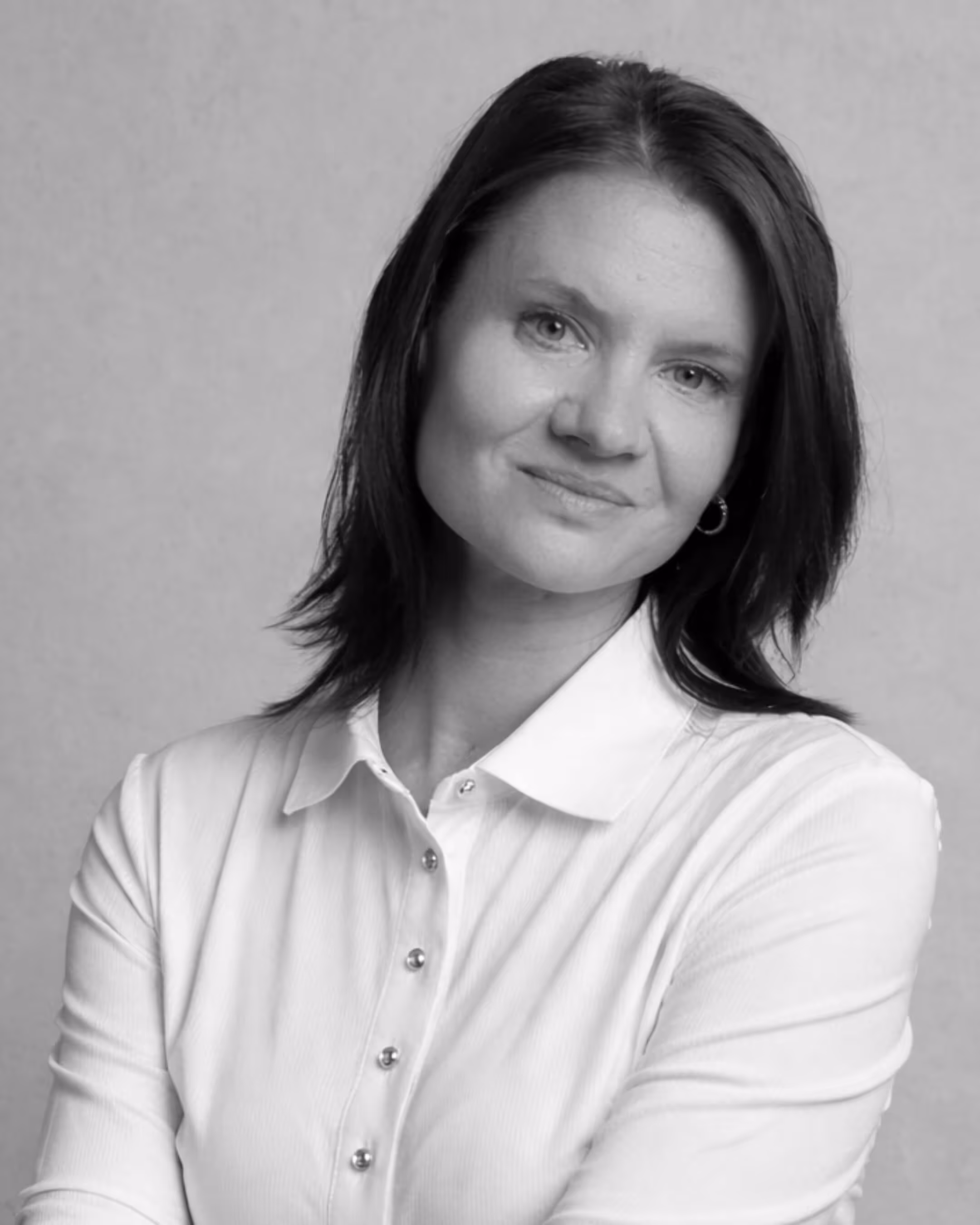 Black and white portrait of a woman with dark hair wearing a white buttoned shirt, smiling slightly with arms crossed.