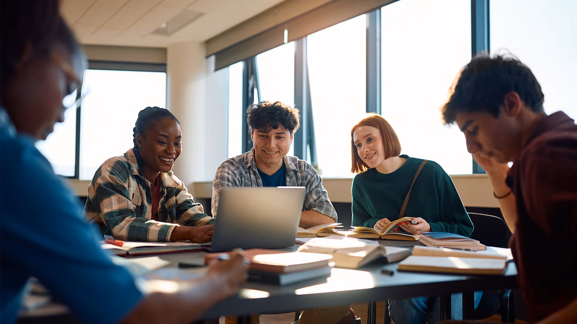 Vier junge Erwachsene lernen gemeinsam an einem Tisch mit Büchern und Laptop in einem hellen Raum mit großen Fenstern.