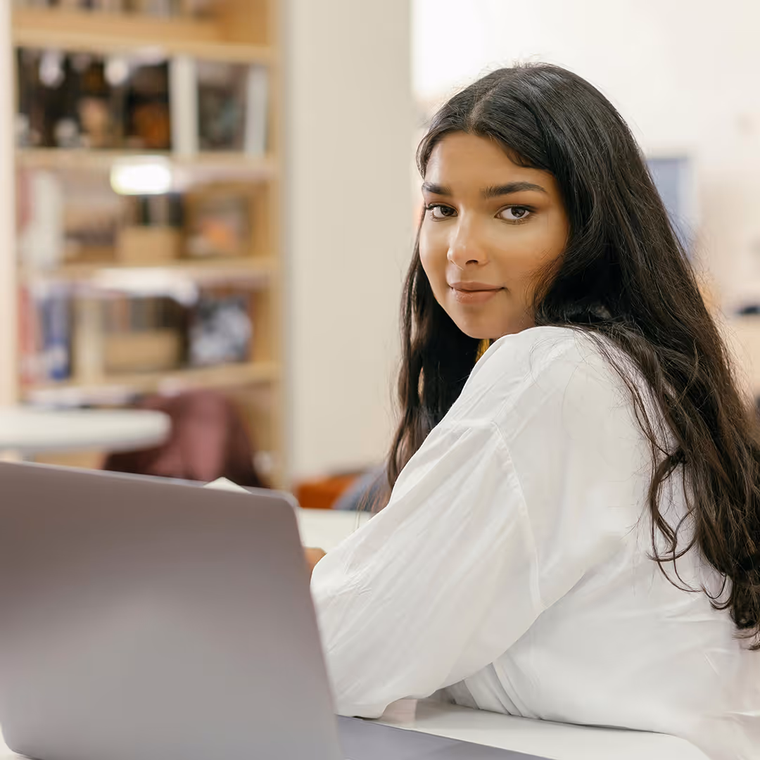 Junge Frau mit langen dunklen Haaren, die an einem Laptop arbeitet und freundlich in die Kamera blickt.