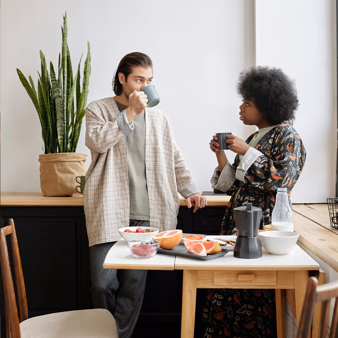 Ein Mann und eine Frau stehen in einer Küche, trinken Kaffee und unterhalten sich, während sie an einem Tisch mit Obst und Küchenutensilien stehen.