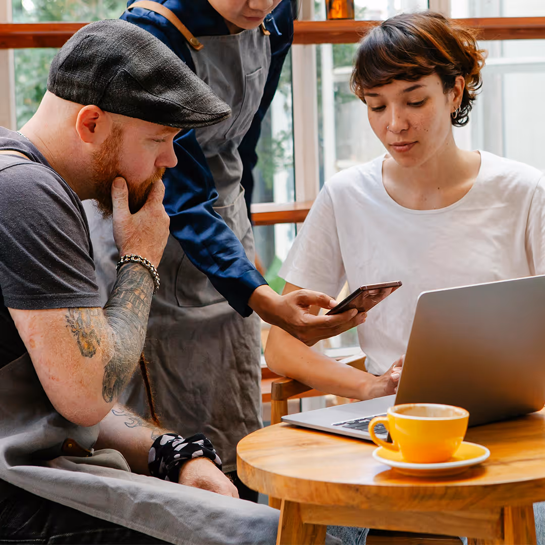 Drei Personen in einem Café, eine sitzt mit Laptop und Hafermilch-Cappuccino, eine andere zeigt ihnen etwas auf einem Smartphone.