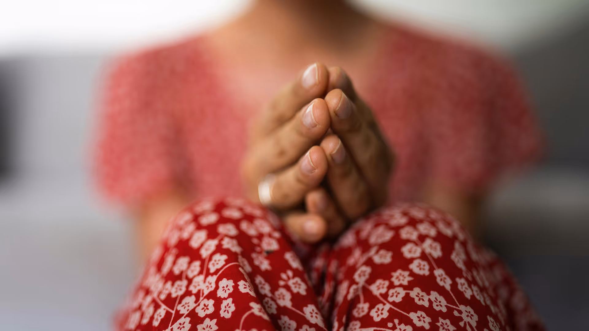 Gekreuzte Hände im Gebet vor Knien in roten, mit weißen Blumen gemusterten Hosen.