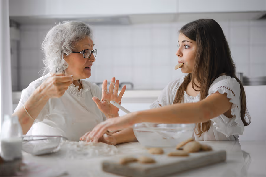Ältere Frau und junges Mädchen backen zusammen Kekse in der Küche, das Mädchen hat einen Keks im Mund.