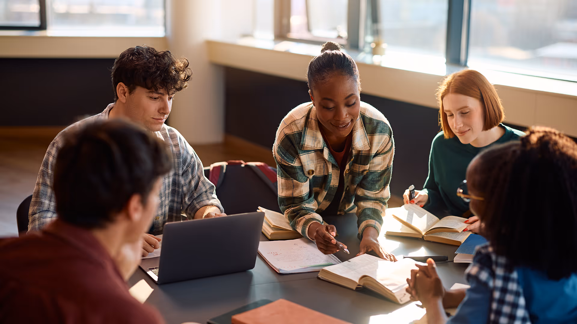 Fünf Studenten sitzen um einen Tisch, lernen gemeinsam und schauen in Bücher und einen Laptop in einem hellen Raum.