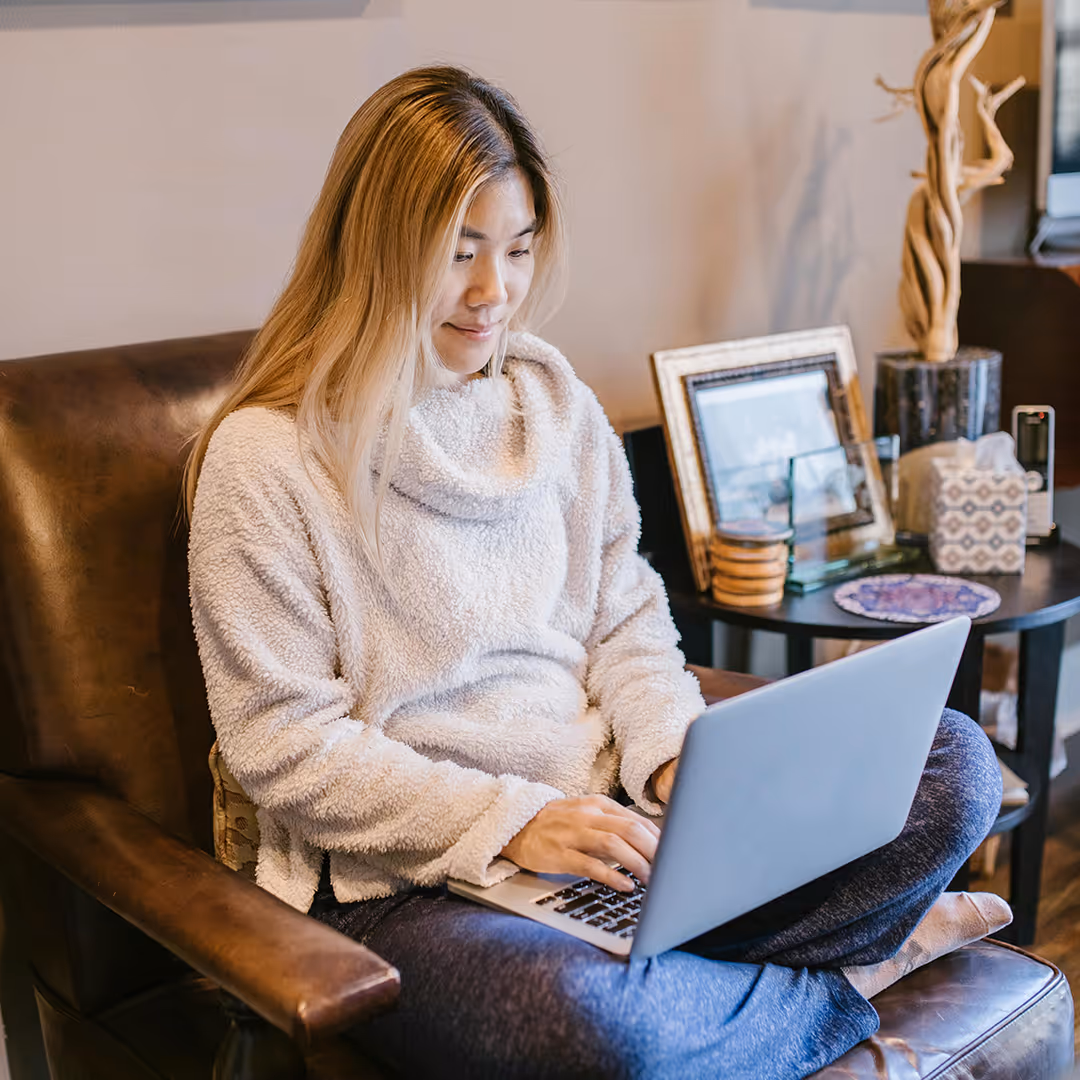 Junge Frau sitzt bequem in einem Ledersessel mit verschränkten Beinen und arbeitet an einem Laptop.