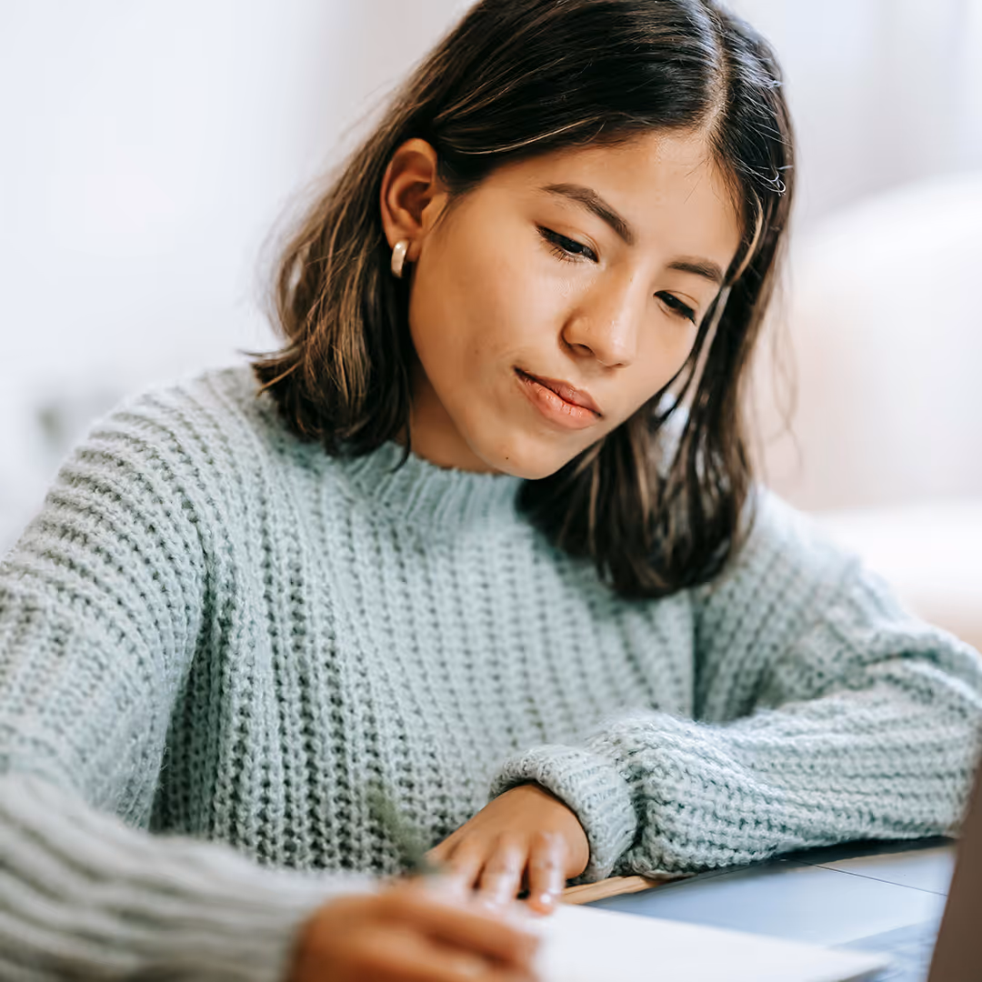 Junge Frau in einem grauen Strickpullover schreibt konzentriert auf ein Blatt Papier, während sie an einem Laptop sitzt.