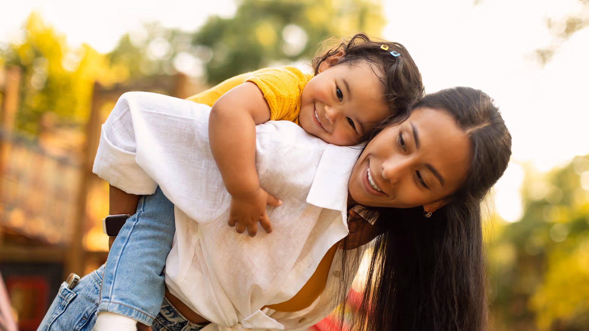 Mutter trägt lachendes kleines Kind auf ihrem Rücken in einem sonnigen Outdoor-Park.
