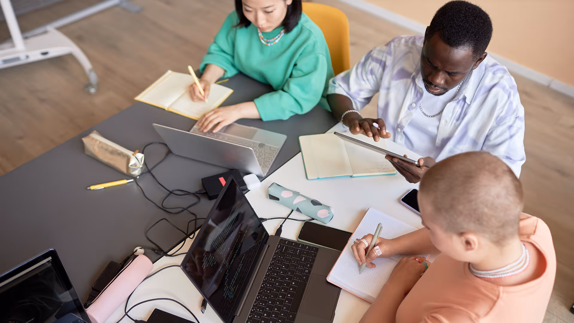 Drei Personen arbeiten zusammen an einem Tisch mit Laptops, Tablets und Notizbüchern in einem modernen Arbeitsumfeld.
