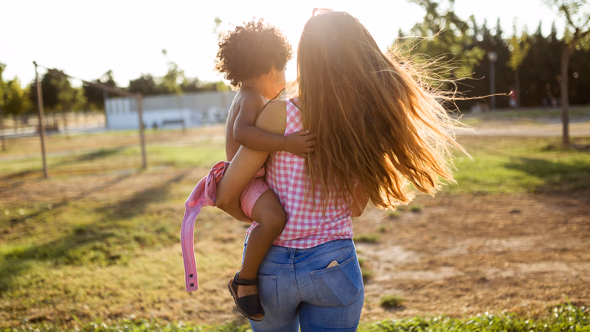 Frau mit langem Haar trägt ein Kleinkind in einem Park bei Sonnenschein.