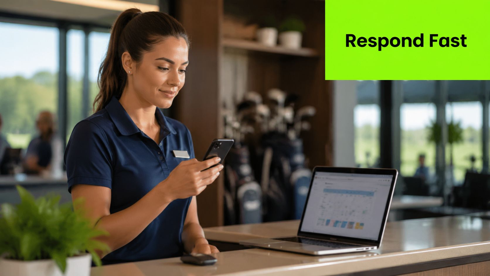 A female staff member at a golf club reception desk using her smartphone while looking at a laptop.