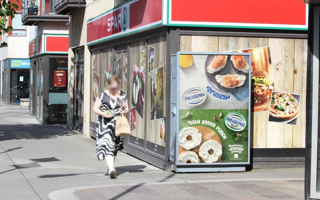 Bauer Media Outdoor digital advertising screen inside a Sainsbury’s supermarket displays a Colgate Max White ad, as shoppers move through grocery aisles.