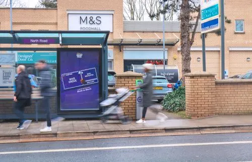 An Adshel bus shelter advertisement on a busy road with people passing by