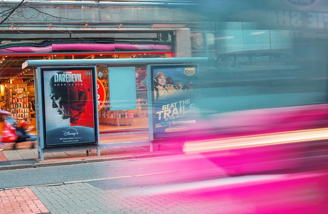 A bus shelter advertisement on a busy street with people walking by and traffic passing