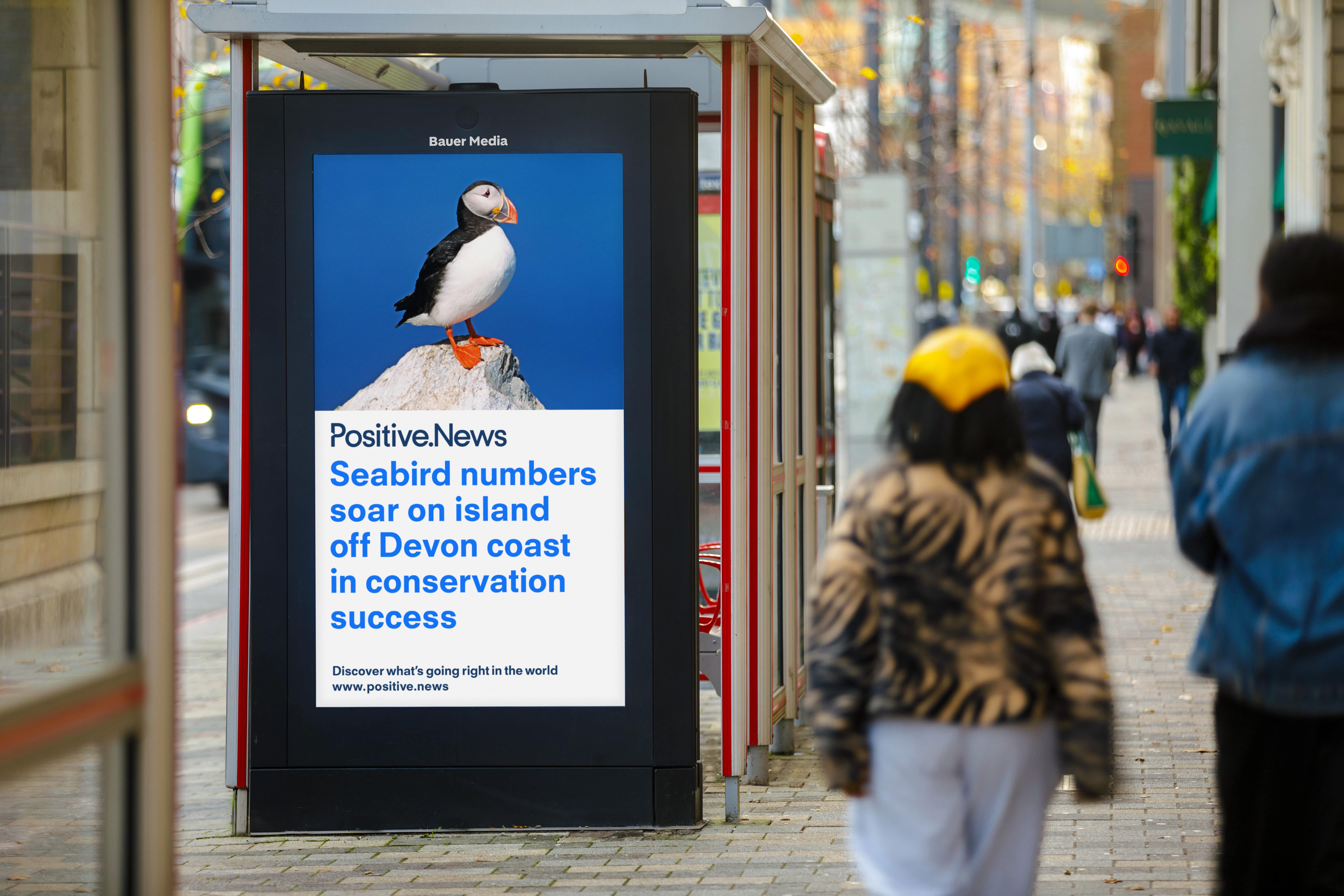 A Positive.News headline displayed on a Bauer Outdoor Media UK Adshel Live display at a bus stop as people walk past during the day