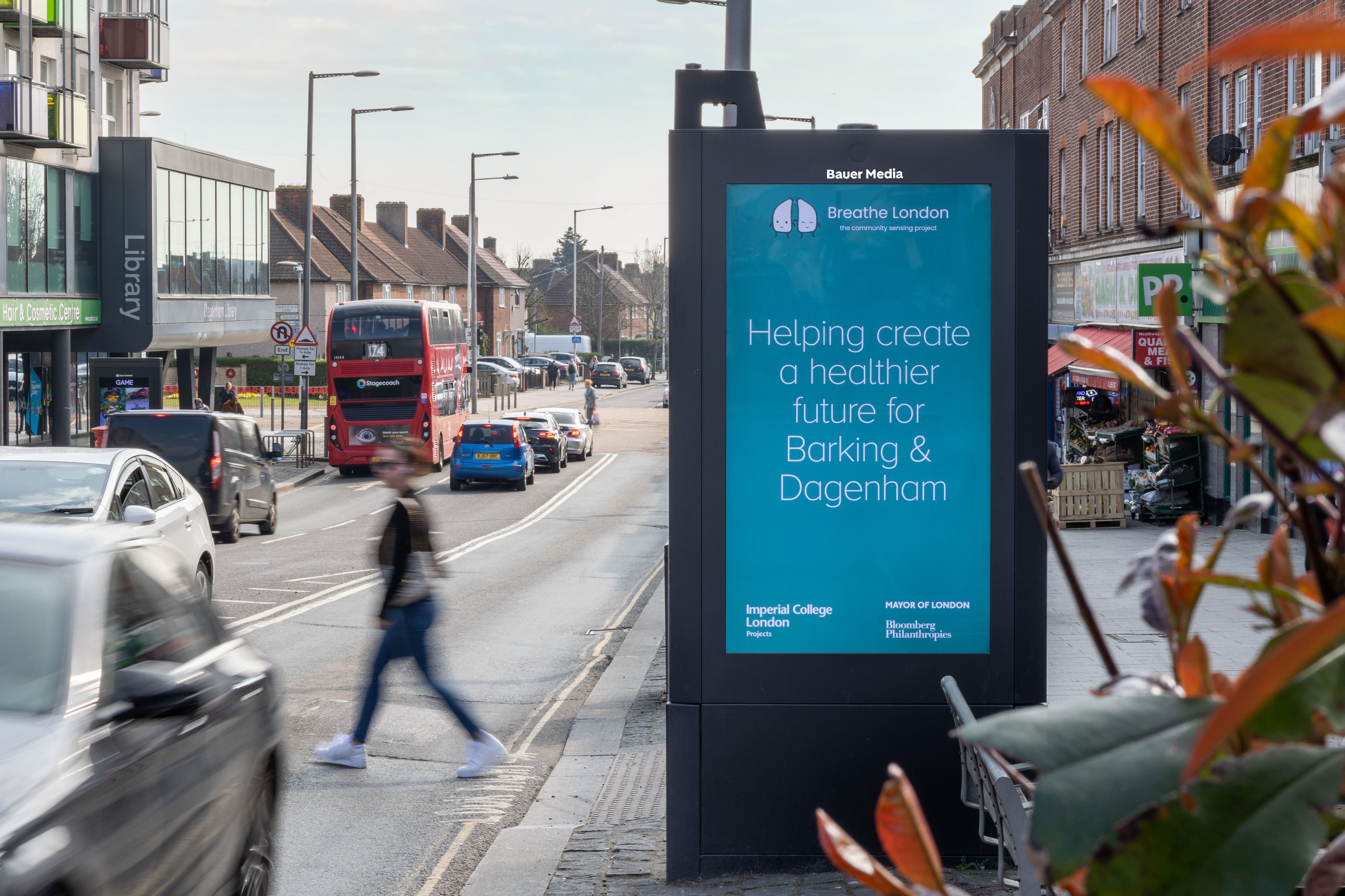 A Breahe London advert features on a digital screen, with an air quality sensor on the top left corner, by a busy road, during the day, as pedestrian pass by
