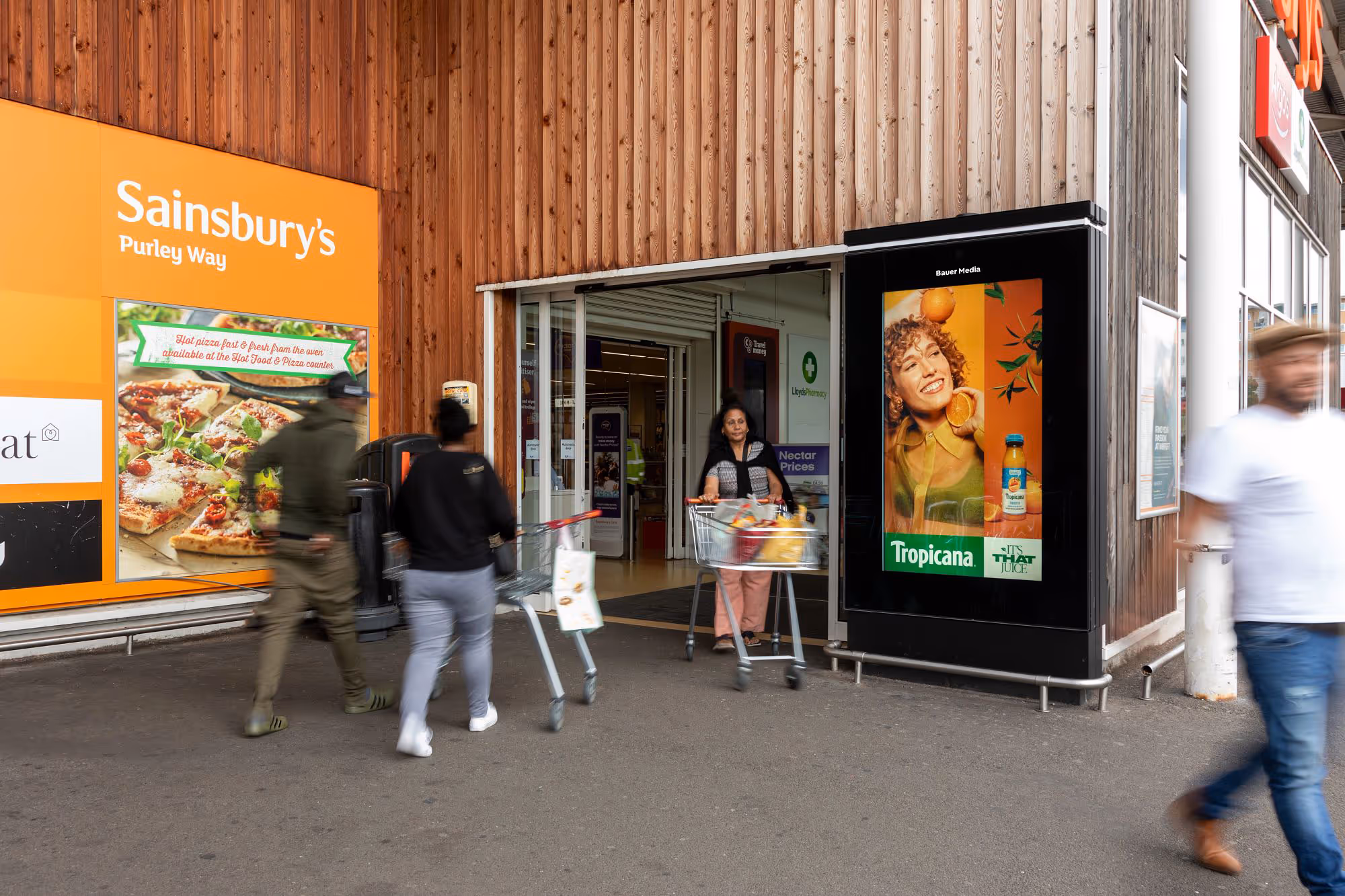 A Tropicana advert features on a digital screen outside a Sainsbury's as shoppers walk in and out of the store, during the day