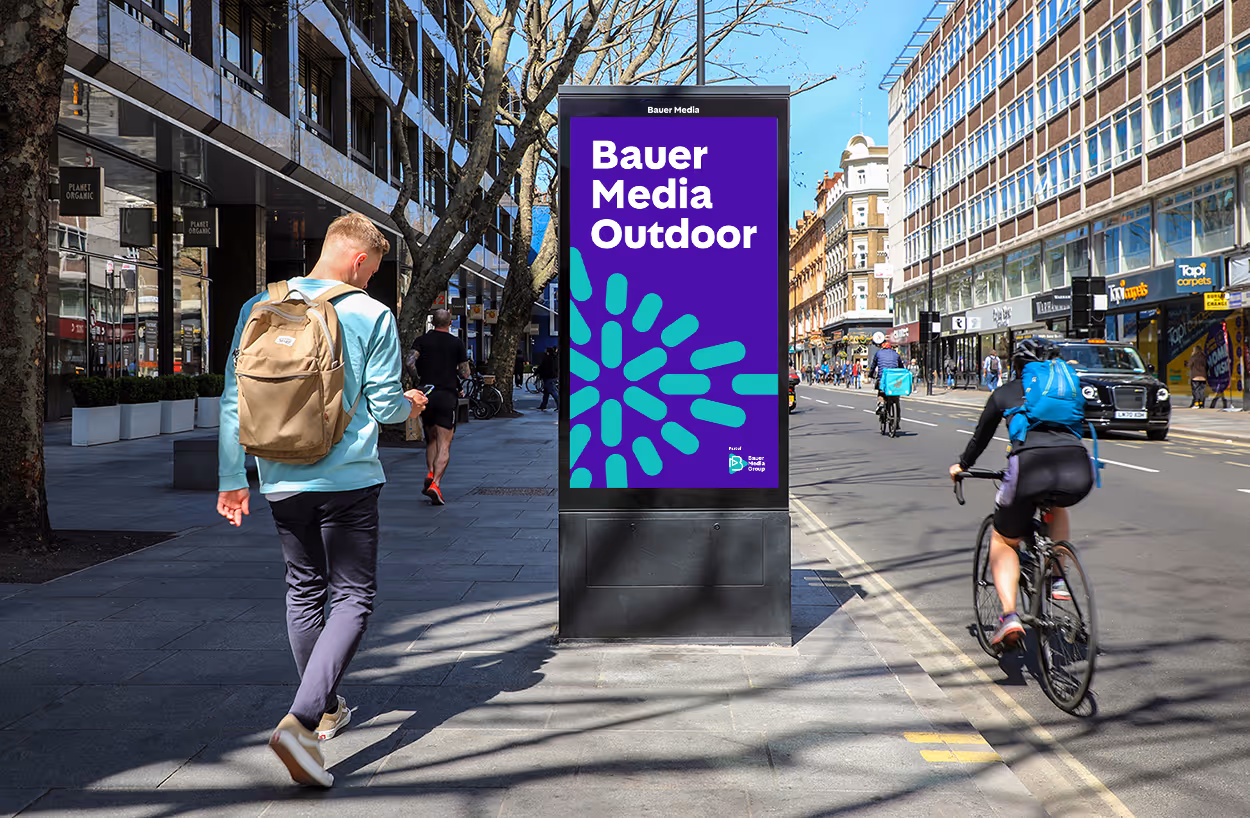 Bauer Media Outdoor logo featured on a digital screen, on a busy street, during the day as pedestrians pass by