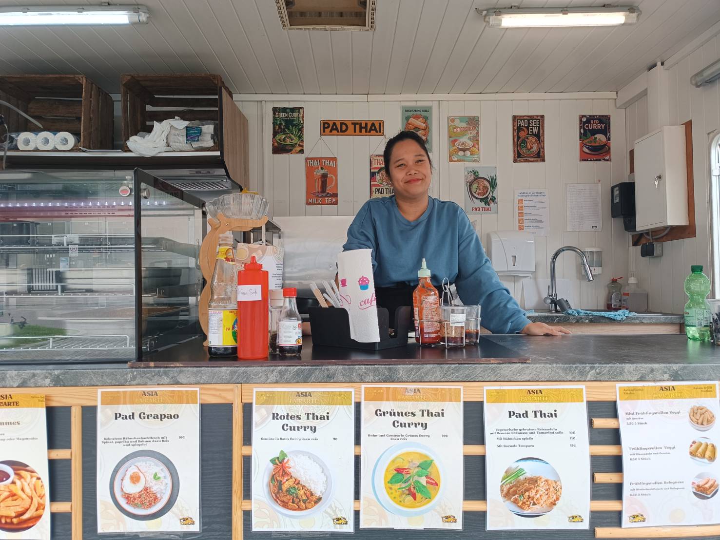 Verkäuferin steht in einem thailändischen Imbiss vor einer Theke mit verschiedenen Saucen und einem Menü mit thailändischen Gerichten.