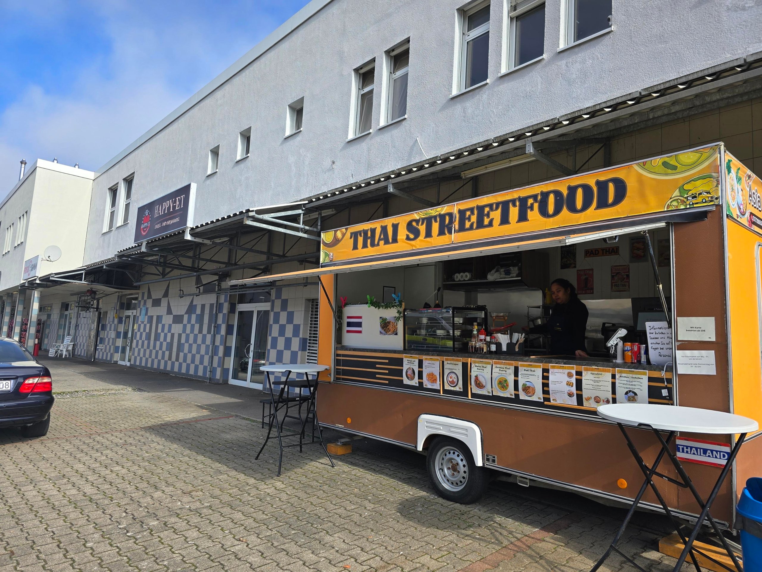 Ein thailändischer Streetfood-Imbisswagen mit gelbem Schild und aufgeklapptem Fenster, davor zwei Stehtische auf einem gepflasterten Gehweg neben einem Gebäude.