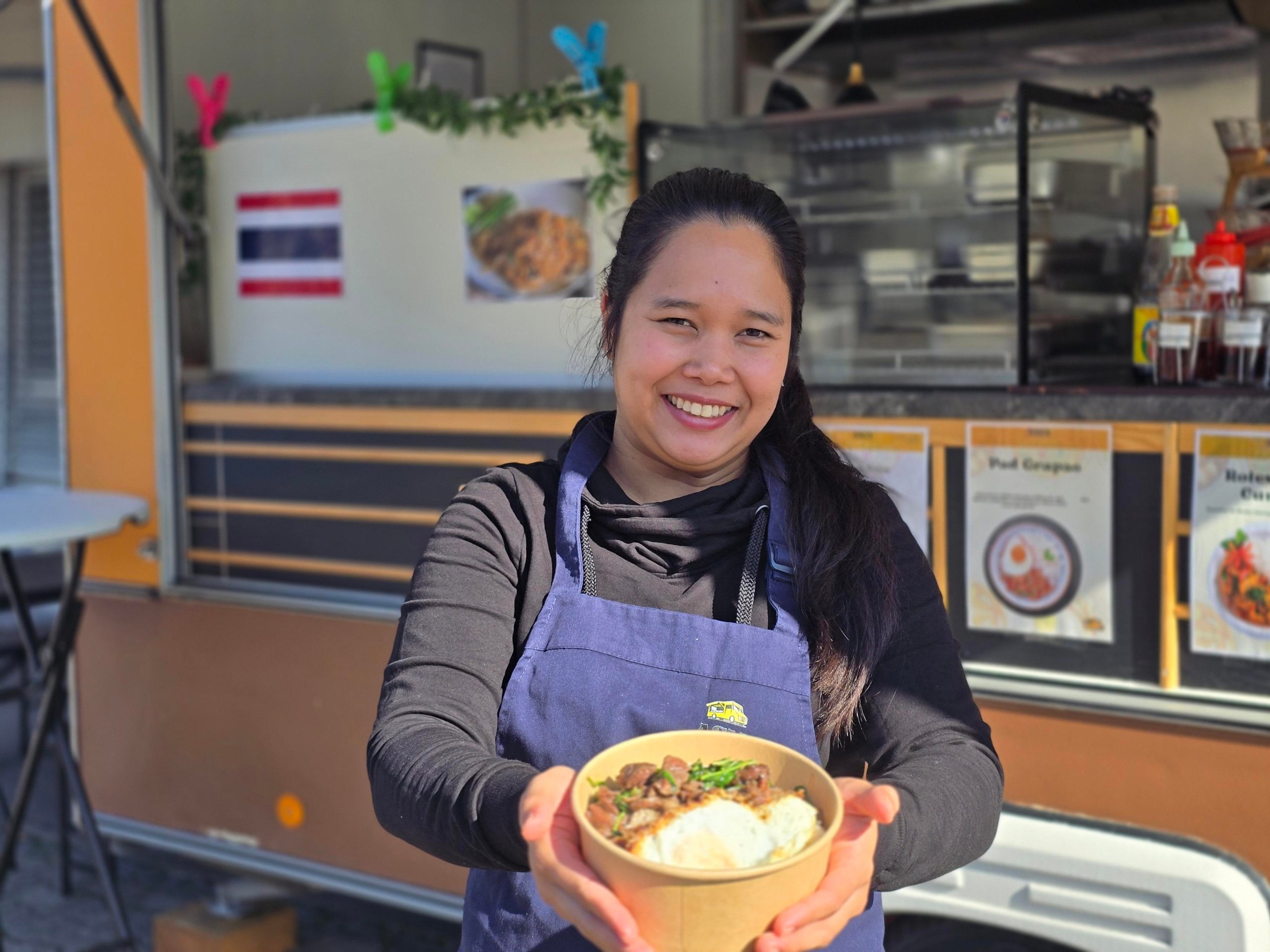 Frau in Schürze reicht lächelnd eine Schale mit Essen, im Hintergrund Imbissstand mit Speisekarten.