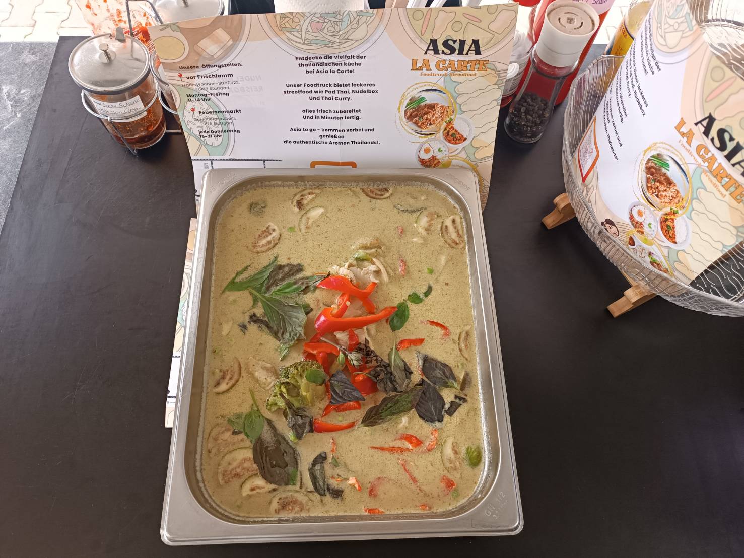 Thailändisches grünes Curry mit roten Paprikastreifen, Basilikumblättern und Pilzen in einem Metallbehälter vor einem Asia La Carte-Menü.