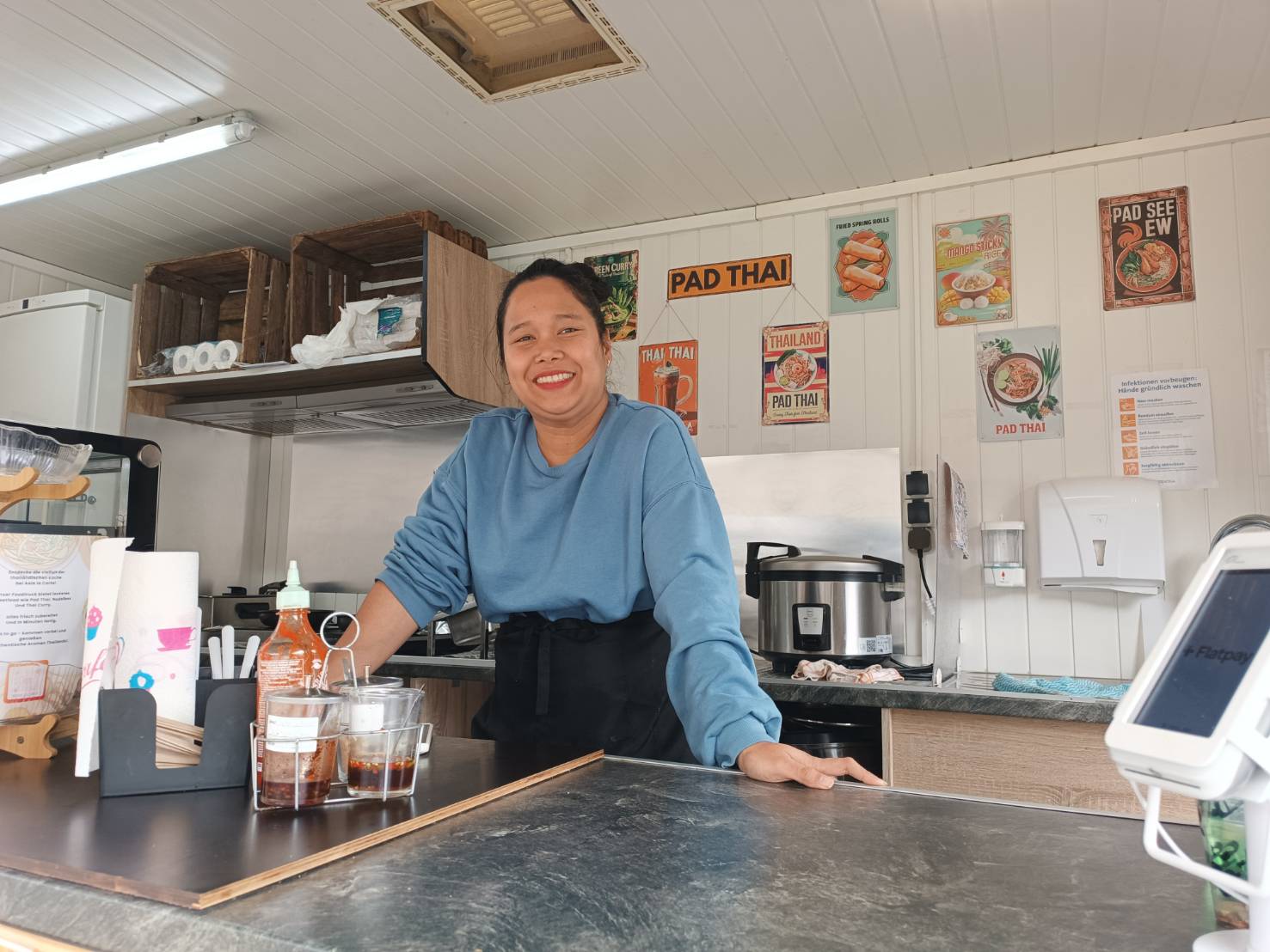 Lächelnde Frau in blauem Pullover steht hinter einem Tresen in einem Thai-Imbiss mit Gewürzen, Reiskocher und Thai-Gerichte-Postern an der Wand.
