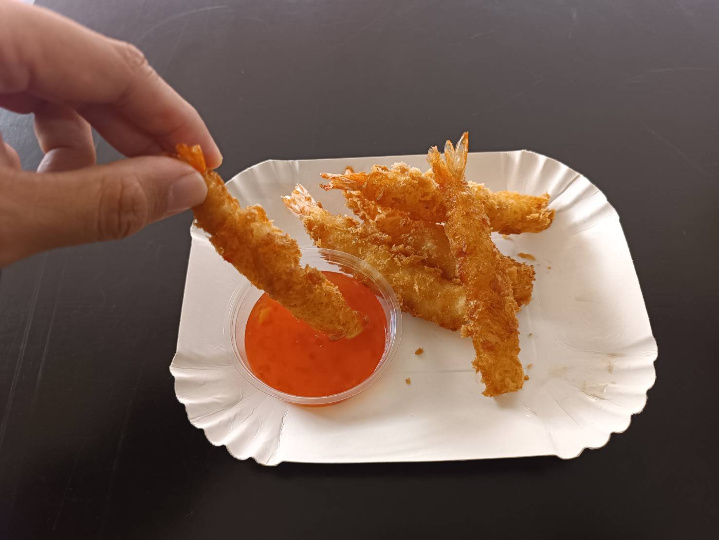 Hand, die ein paniertes gebackenes Garnelenstück in eine Schale mit roter Soße taucht, auf einem Papierteller mit weiteren Garnelen.