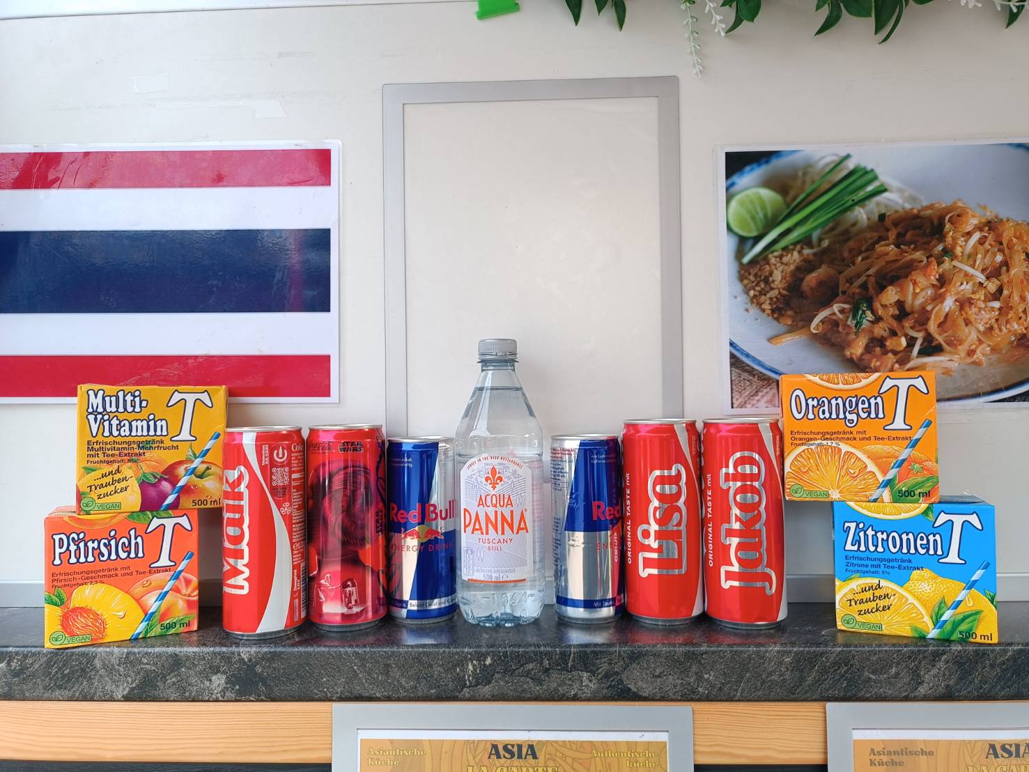 Verschiedene Getränkedosen und -kartons, darunter Red Bull, Coca-Cola und lokale Marken, auf einer Theke vor einer thailändischen Flagge und einem Teller mit asiatischem Essen.