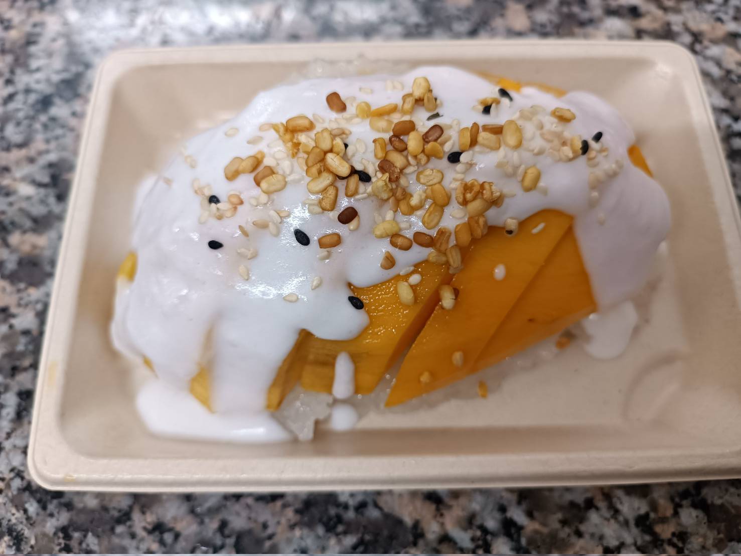 Geschnittene Mangoscheiben mit Kokossahne und Sesamkörnern auf einer rechteckigen beige-farbenen Platte.