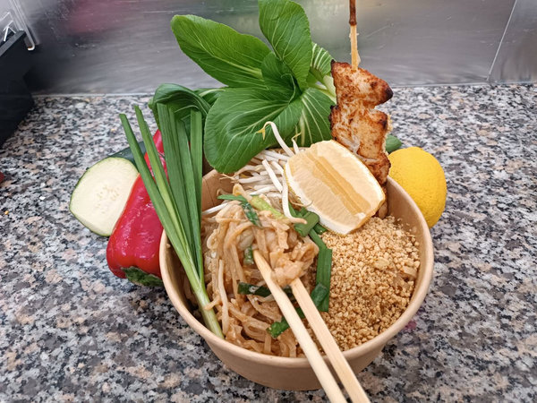 Schale mit Pad Thai, garniert mit Erdnüssen, Zitronenscheibe, Sprossen, frischem Gemüse und gegrilltem Hähnchenspieß, Essstäbchen halten Nudeln.