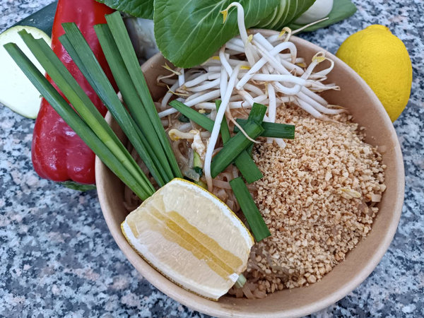 Schüssel mit Pad Thai, garniert mit Erdnüssen, Limettenscheibe, Frühlingszwiebeln und Bohnensprossen, umgeben von frischem Gemüse und Zitrone auf Marmoroberfläche.