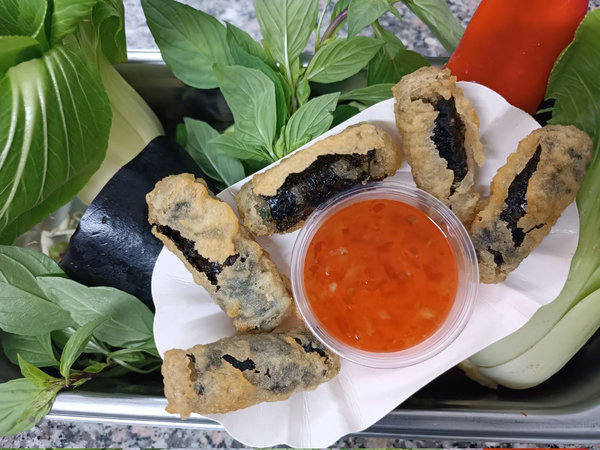 Fünf frittierte Frühlingsrollen auf einem weißen Teller mit einer Schale süßer Chilisauce, umgeben von frischen Kräutern und Gemüse.