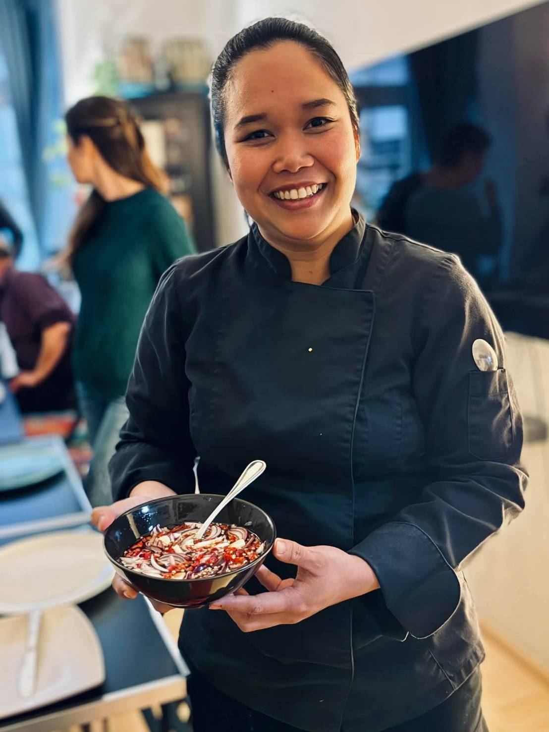Lächende Köchin in schwarzer Kochjacke hält eine Schüssel mit Essen und einem Löffel darin.