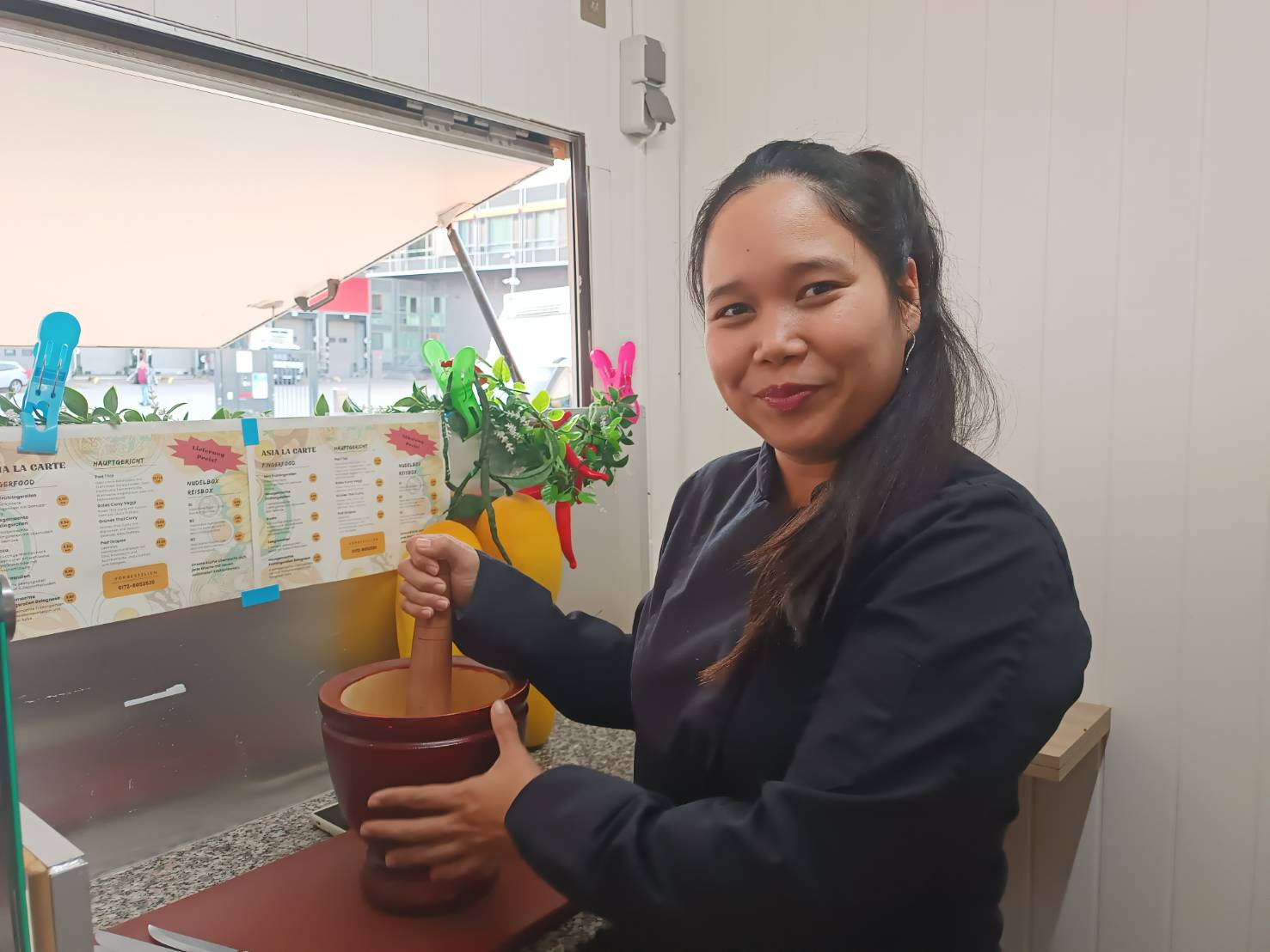 Frau mit dunklen Haaren bindet ihre Haare zurück und benutzt einen Mörser und Stößel in einem Fensterbereich eines Restaurants.