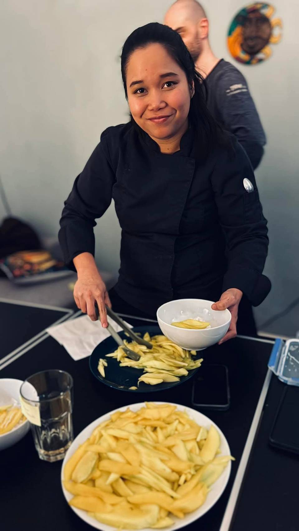 Köchin in schwarzer Jacke hält eine weiße Schüssel mit Sauce und verwendet Zangen, um Pommes frites auf einen Teller zu legen.