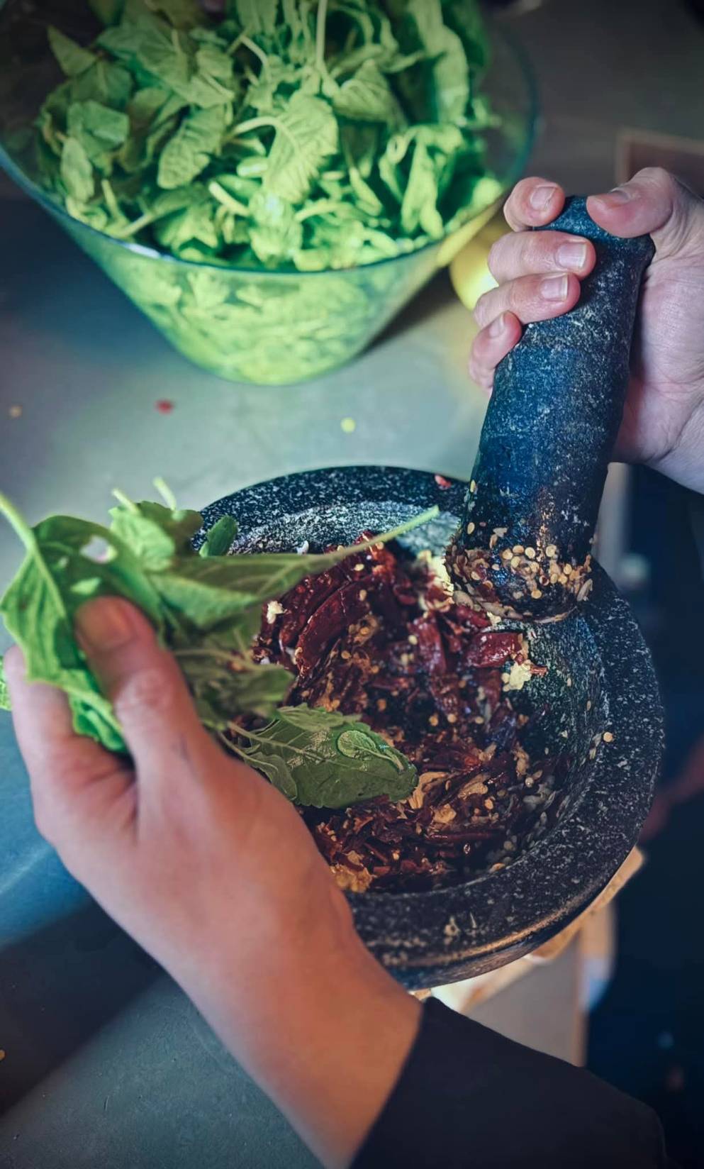 Person zerkleinert getrocknete Chilischoten mit einem Mörser und Stößel, hält frische grüne Blätter bereit.