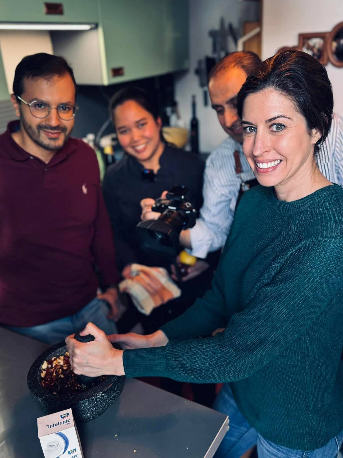 Frau mahlt Zutaten in einem Mörser, während drei Menschen im Hintergrund lächelnd zuschauen, eine Person hält eine Kamera.