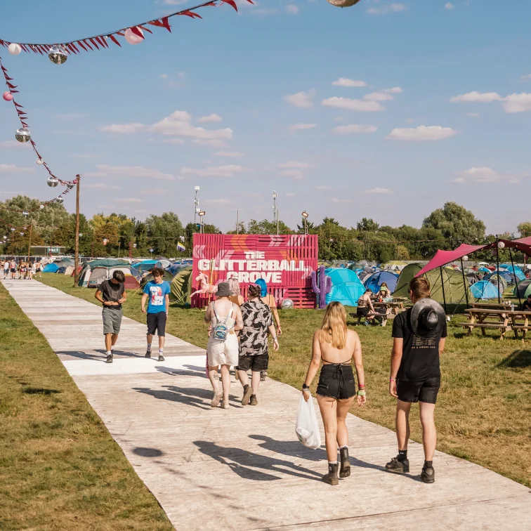 Festivalgoers walking toward The Glitterball Grove area with tents, flags, and disco balls under a blue sky.