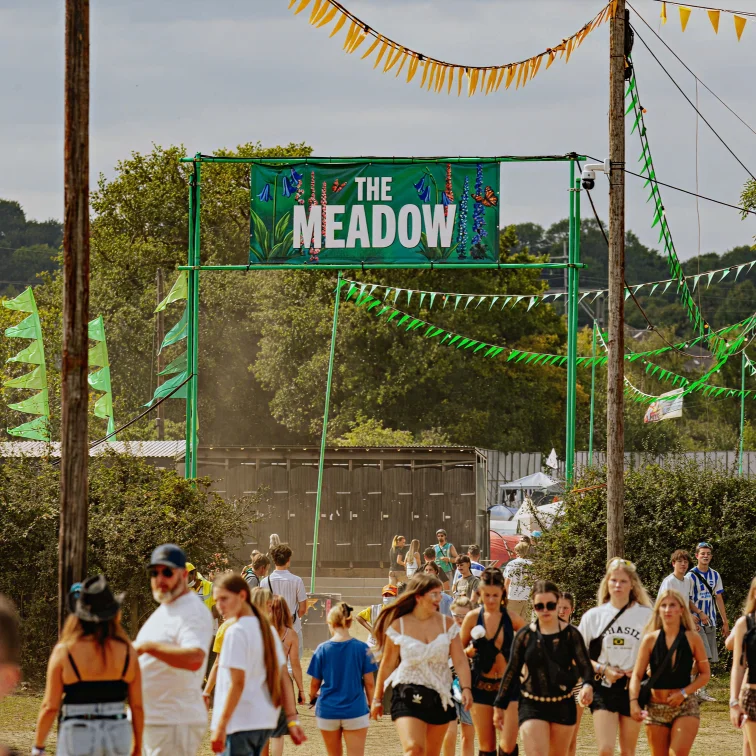Crowd of festival attendees walking beneath green bunting toward The Meadow entrance on a sunny day.
