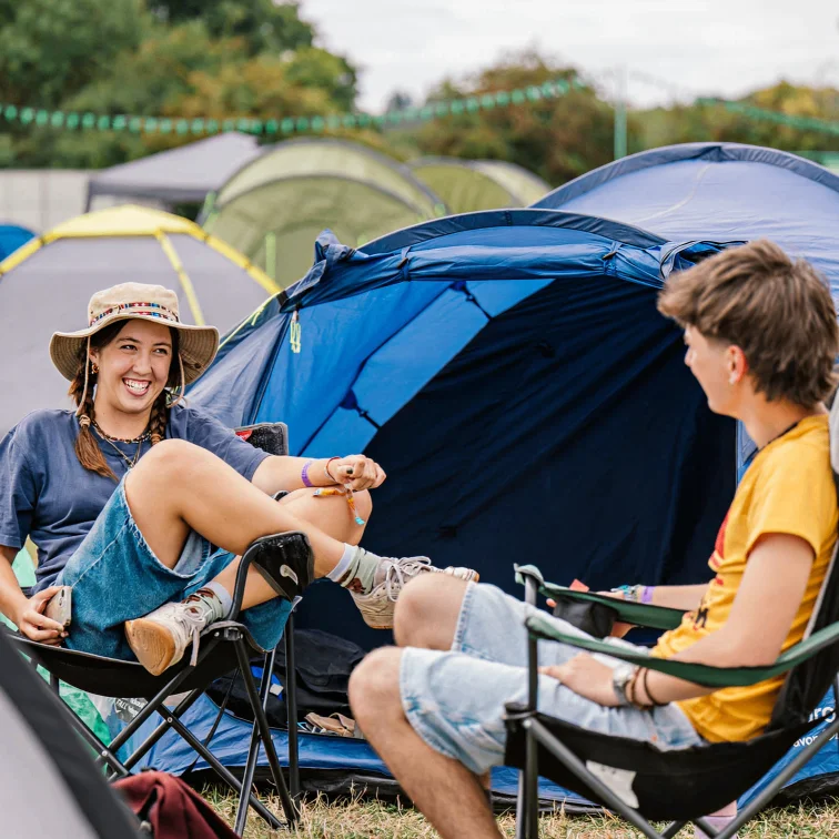Two friends relaxing on camping chairs beside blue tents at a festival campsite, smiling in the sun.