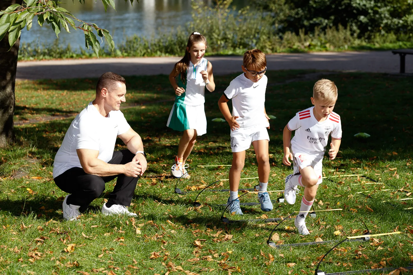 Ein Mann in weißem T-Shirt kniet im Gras und trainiert drei Kinder, die eine Hindernisleiter im Park überspringen.