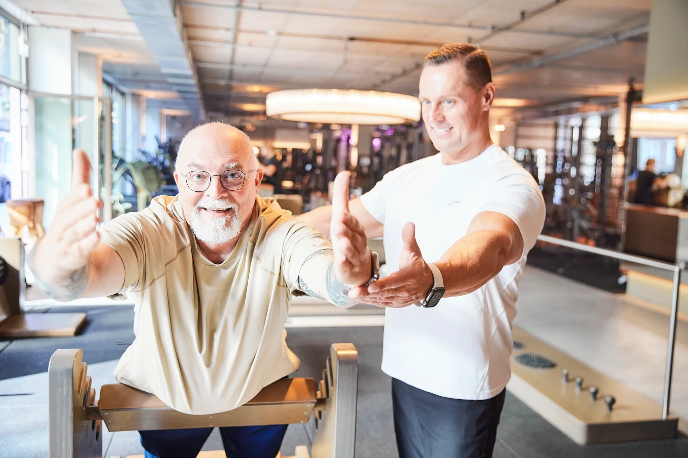 Älterer Mann trainiert mit Trainer im Fitnessstudio, beide strecken die Arme nach vorne.