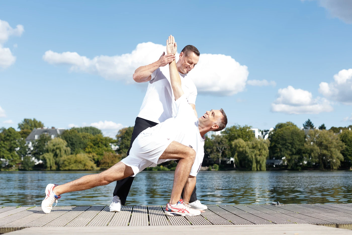 Zwei Männer tragen beim Training draußen am Wasser eine Dehnübung mit Handklatschen aus.