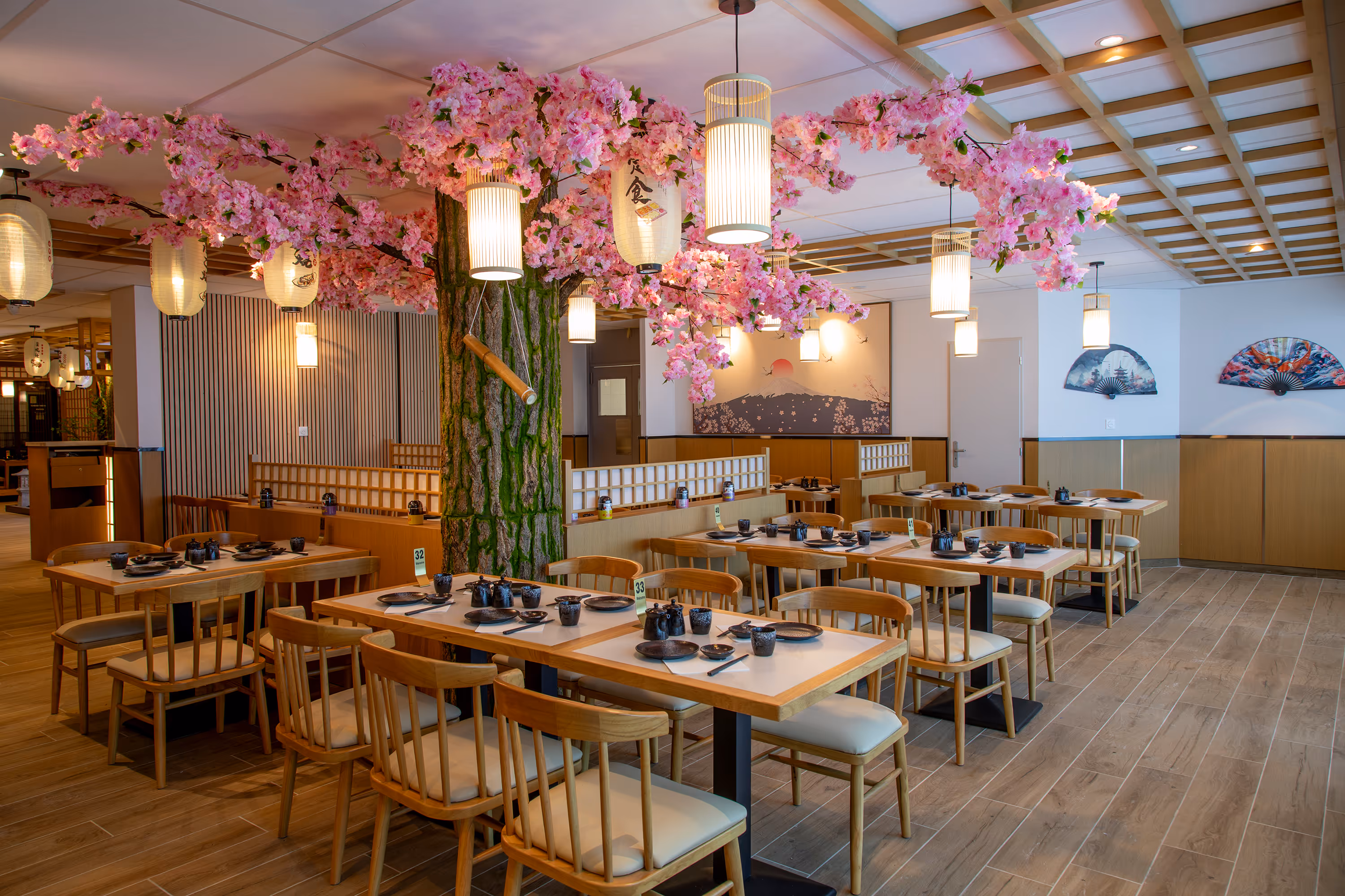 Intérieur d'un restaurant japonais avec tables en bois, décorations florales roses suspendues simulant un arbre, lanternes traditionnelles et éventails décoratifs au mur.