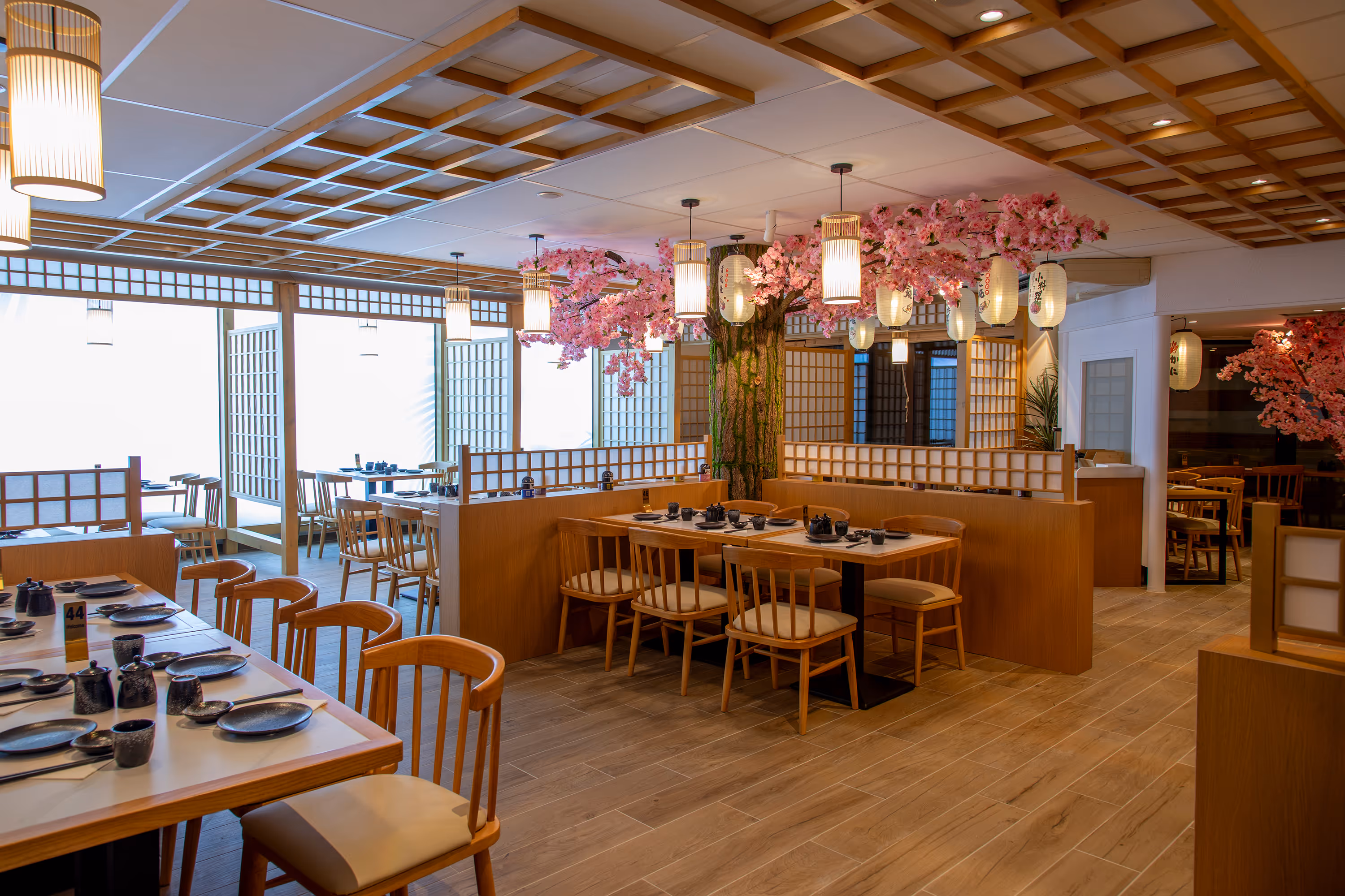 Intérieur d'un restaurant japonais avec des tables en bois, lanternes suspendues, et arbres de cerisier en fleurs décoratifs.
